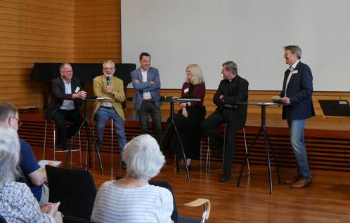 Vier Männer und eine Frau stehen an Stehtischen in der Aula und diskutieren.