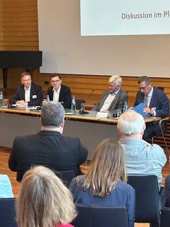 Vier Personen sitzen am Tisch in der Aula, im Vordergrund sind einige sitzende Teilnehmende von hinten zu sehen.