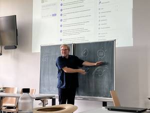 Ein Mann in dunkelblauer Kleidung und grauen Haaren lächelt in die Kamera und erklärt den Teilnehmenden eines Seminars etwas an der Tafel, über der Tafel erscheint die digitale Präsentation.
