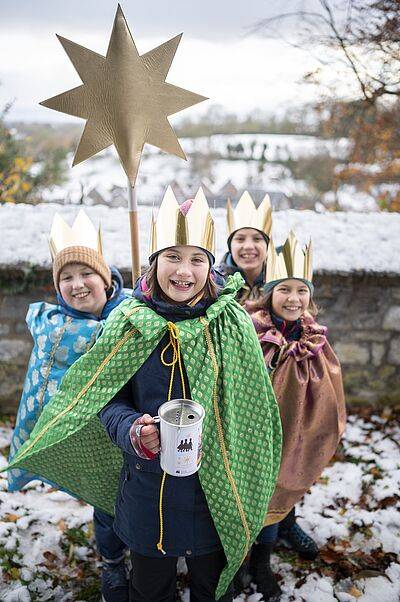 Vier Kinder mit goldenen Kronen und bunten Umhängen stehen im Schnee, eines hält einen großen goldenen Stern auf einer Stange und eine Spendendose