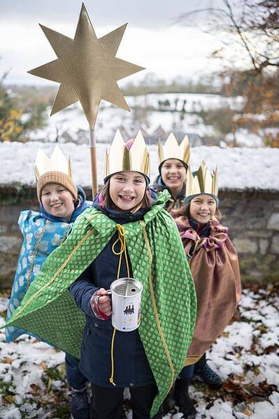 Vier Kinder mit goldenen Kronen und bunten Umhängen stehen im Schnee, eines hält einen großen goldenen Stern auf einer Stange und eine Spendendose